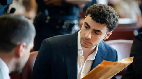 Luigi Mangione looks toward his lawyer while wearing a navy jacket and white dress shirt. He is holding a yellow envelope in his hand.