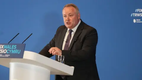 Darren Millar speaking at a podium which says Fix Wales and Trwsio Cymru. He is wearing a black suit, dark tie and white shirt.
