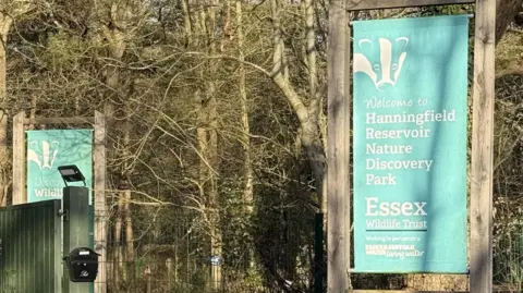 A blue sign for the reservoir. There are several bare trees behind the sign.