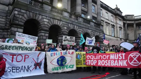 PA Media A group of protesters from climate organisations outside the Court of Session in Edinburgh. From left to right, one group are holding a greenpeace banner, which has the name of the organisation in green on a white banner. Below that is the words "Stop Rosebank" with stop in red and Rosebank in black. An image of an oil platform with a red X over the top is next to it. The next banner reads "Edinburgh" in multi-coloured letters and Greenpeace in green on a light-coloured banner either side of a globe in blue and green with a red eye shape surrounding it. The final banner is red with the words "equinor must stop Rosebank" in white letters.