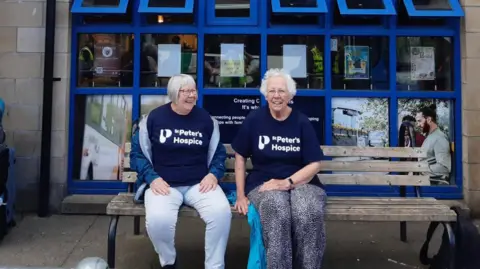 Valerie Boon The two women sat on a public bench in Inverness waiting for a bus. They are both sat down and smiling at the camera. They are wearing blue charity t-shirts.