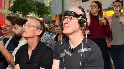 Prof Damian Bailey sits beside James Gallagher, who is wearing a head-mounted device with wires and sensors attached, as they watch football on TV in a packed pub