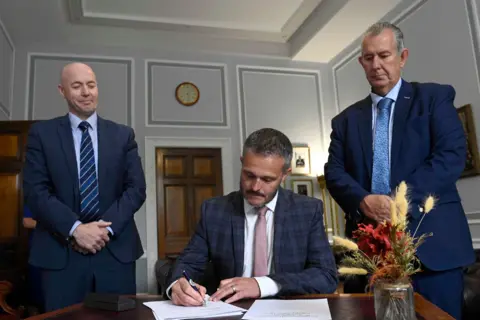 Mark Marlow/PA Wire Ulster Unionist leader Jon Burrows (left) and Assembly Speaker Edwin Poots MLA (right) watch as Ulster Unionist MLA Robbie Butler signs the pledge of office at Stormont Buildings, Belfast, following his appointed as new Health Minister.