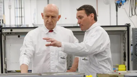 Reuters Defence Minister John Healey speaks to a man on an assembly line at a factory in Stevenage. Healey is bald and has blue eyes.
