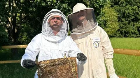 David Maxen David Maxen smiling for a photo with a friend of his who is holding a super covered with bees. They are wearing a white and a beige beekeeping suits. It is a sunny day.