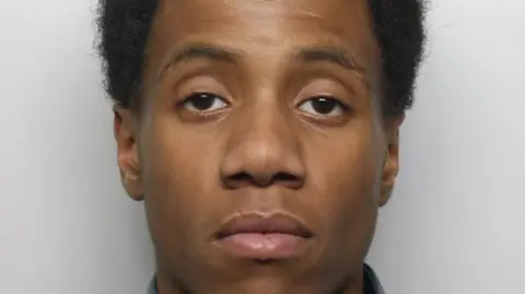 Romaine James' mugshot, where he is looking at the camera with a serious expression in front of a grey background. He has black hair.