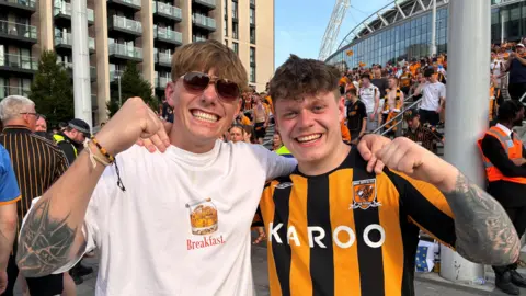 BBC/ Linsey Smith Two young men hold their fists aloft and smile, the man on the left is wearing sunglasses, then man on the right is wearing a Hull City football shirt.