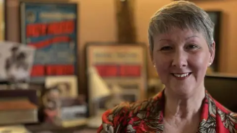 Debbie Hudd smiles at the camera. She has short grey hair and wears a red blouse with black and white flowers on it. 