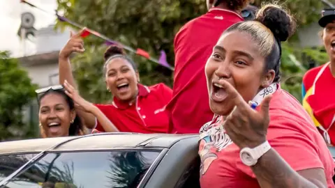 Three women in red T-shirts smiling and waving their hands in the air in celebration as they lean out of a car.