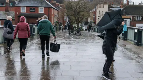 Getty Images Four people walking over a bridge in the rain. On the right hand side, someone is struggling with an umbrella that has turned inside out in the wind.