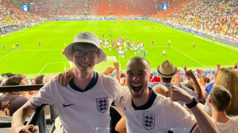 Ben Price Ben right hand side wearing a white football shirt and is posing with his friend who is wearing a bucket hat behind a football staduim backdrop. 