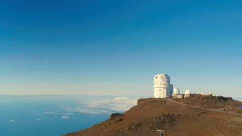 NSF National Solar Observatory The image shows an observatory - a white, dome-like structure, perched on a peak overlooking the ocean. Scattered clouds are visible below the observatory and the sky above is clear and blue. This is the NSF Inouye Solar Telescope - the most powerful solar telescope in the world.