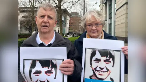 A man and a woman holding black and white pictures of a teenage boy. They are standing outside. 