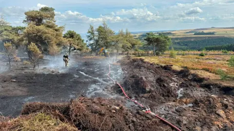 A moorland field has been half burned away with just blackened soil. In the distance is a firefighter and a tractor by some trees