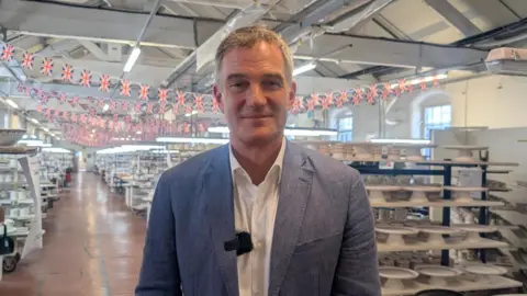 Peter Kyle wears a blue jacket and white shirt, he is standing at Emma Bridgewater factory, surrounded by tableware and union jacks. 