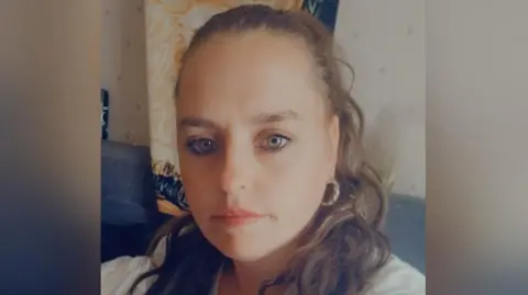 Ms Blackham is smiling in a selfie photo. She has brown hair tied back and is wearing a white top and is sat inside a home.