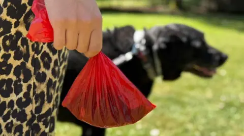 A close-up of a woman's hand holding a red poo bag. She wears leopard print trousers. A dog can be seen in the background, standing in a grassy field.