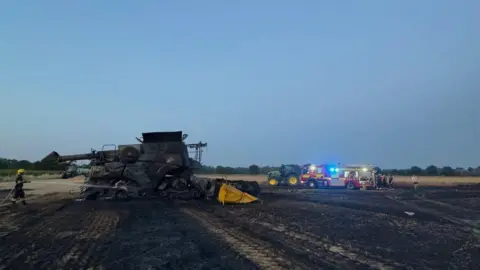 Cambridgeshire Fire and Rescue Service The blackened remnants of a combine harvester surrounded by a burned field. One firefighter is hosing the machinery down, and in the distance is a fire engine surrounded by people