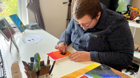 a man sat at a table painting. He had a dark top on, glasses and brown hair.