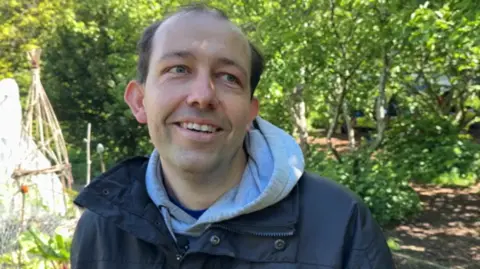 Keith Bosomworth, also a volunteer - also smiling. He is a man in his mid-30s with thinning hair and wearing a jacket with a hoodie underneath. To one side behind him is a small allotment. Bamboo sticks to support the growing fruit or vegetables are pointing into the air.