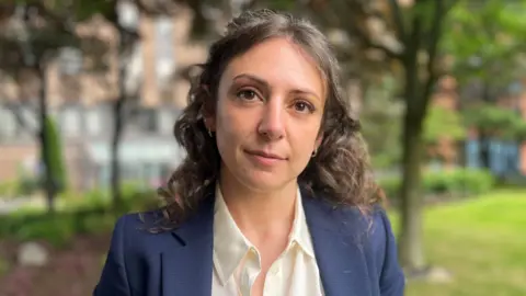 Allison Catalano Head-and-shoulders outdoor portrait of a person with shoulder-length curly hair, wearing a navy blazer over a white shirt. Trees, grass and buildings are visible in the softly blurred background.