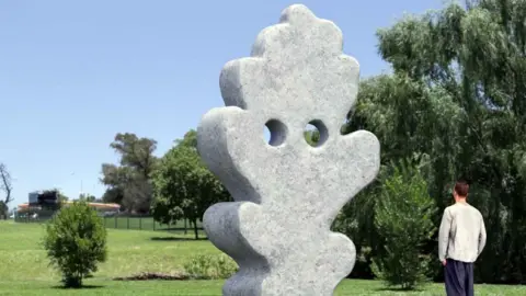 A computer generated image of what the installation will look like. It is a large, 13 foot tall sculpture made of stone. It is shaped irregularly like a leaf, with two large holes towards the top. A man is stood on the right, admiring the sculpture. It is a sunny day and there are lots of mature trees in the background.