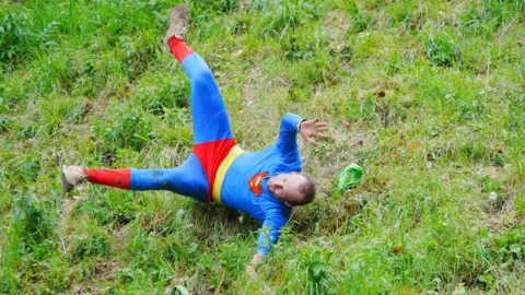 Ben Birchall/PA Wire A man in a blue and red Superman costume tumbling sideways down a grassy hill. His legs and arms are stretched out wide and he is wearing a focused expression.