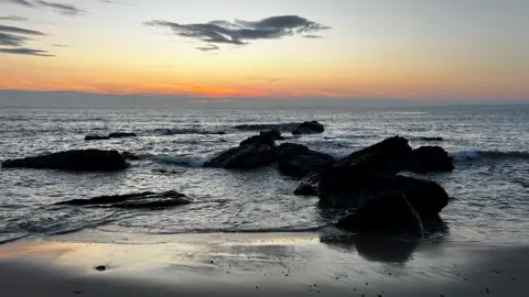 Catherine Ronald A view of rocks jutting out of water against and orange and yellow evening sky