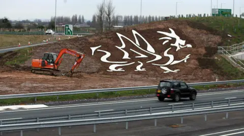 The eastbound side of the A494 dual-carriageway in Flintshire showing work being completed on a white dragon shape cut into a sloping embankment. The design is filled with white stone so that it stands out to passing motorists. A digger can be seen on the muddy ground.