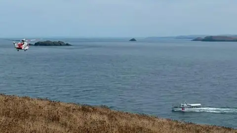 HM Coastguard Padstow The rescue helicopter is descending on the bay and a white leisure boat with canopy is sailing to the same point. It is a clear blue sky day and the sea is rippled. There are two rocky islands and the coastline can be seen receding to the horizon. The grass at the top of they cliff in the foreground is brown.