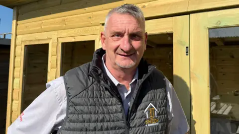 Grace McGrory/BBC A man with grey hair wearing a gilet with a company logo on it. He is standing in front of a wooden summer house.