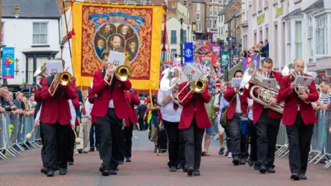 The Durham Miners' Gala. A brass band is walking down the street in red blazers. A red, yellow and blue miners' banner with five men depicted in the middle is being held up behind them. More banners and people can be seen walking behind them with people behind barriers on either side of the street.