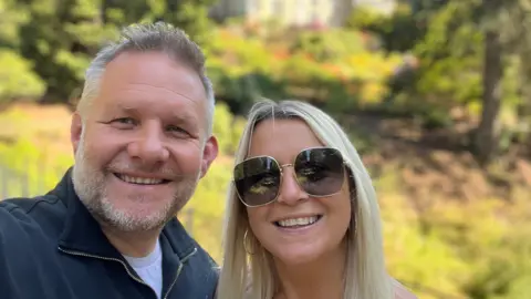 Ben Giles Selfie photograph of Ben Giles (left) and his wife Lindsey(right). Ben has grey short hair, a grey beard, and blue eyes. She smiles widely at the camera. Lindsey wears big wide sunglasses with a gold frame, gold hoop earrings and smiles at the camera. She has bright blonde long hair. Behind them is green grass highlighted by the sun, some trees and a manor house.