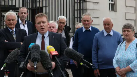 PA Media Solicitor Ciaran O'Hare is speaking to the media outside court. He has brown hair and is wearing a suit. He is surrounded by microphones and is flanked by a group of six men and two women.