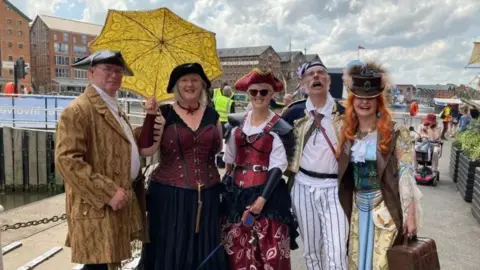 BBC A group of five at the Tall Ships Festival 2024. There are two men and three women. They are all dressed up as pirates and smiling at the camera. 