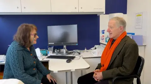 Nicola Haseler/BBC Dr Karen Benson sits in her clinic room within a pharmacy in Letchworth in Hertfordshire. She is wearing a chunky blue/green cardigan with wooden buttons up the front. It has a collar. She has glasses on and has brown shoulder length wavy hair. There is a computer in the centre of the doctor and the patient. She is leaning forwards as if she is paying attention to him. He has grey hair and a bright orange scarf. The patient also has a dark brown tweed jacket