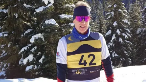 Rob Shenton A man wearing skiing attire while standing on snow-covered ground with trees in the background. He is wearing pink sunglasses and a yellow jacket that has the number 42 on it.