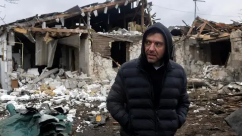 Reuters A man wearing a dark puffer jacket and hoody stands in front of a house that has been damaged by an attack, with the roof blown out and bricks scattered all over
