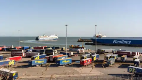 Getty Images Truck trailers parked dockside at Rosslare Europort in Rosslare, Ireland. There is a ferry in the sea in the distance and blue sky.