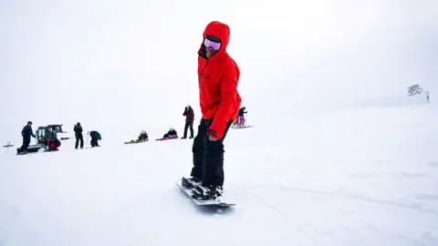 A boarder in a red jacket and black trousers on a board in snow at Cairngorm Mountain.