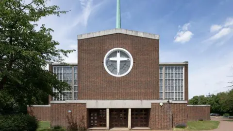 The Roman Catholic Church of Our Lady of Fatima in Harlow, Essex. It has a brick built entry risking to a peaked roof with a slim spire, beneath which is a circular window with a white cross. On either side are two wings, set back, which are entirely glazed