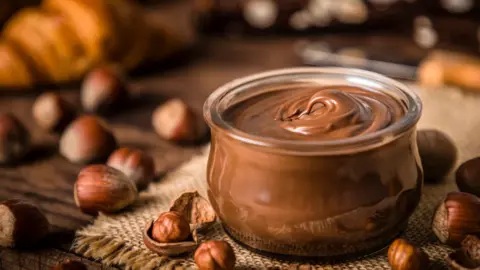 A jar of chocolate and hazelnut spread surrounded by hazelnuts