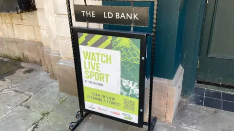 Alex Pope/BBC A sign outside a pub that reads "Watch live sport", in front of a wall.