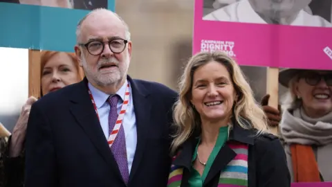 PA Media Lord Falconer and Kim Leadbeater stood next to each other at a protest. Lord Falconer is on the left and is wearing a black suit, purple tie and blue shirt, and black rimmed glasses with a beard. Kim Leadbeater is wearing a green shirt, black jacket, and a multicoloured striped scarf.