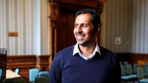 REUTERS/Russell Cheyne Head of Burnley Council Afrasiab Anwar is photographed in the Town Hall in Burnley. He is smiling and looking to his right hand side. He is wearing a white open necked shirt under a navy V-necked jumper.