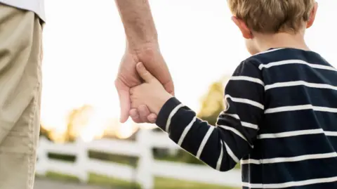 Tampilan belakang Getty dari ayah dan anak berpegangan tangan sambil berdiri di luar ruangan