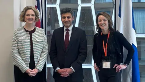 A man in a dark suit stands between two women. The woman on the left is wearing a dark skirt and light patterned jacket. The woman on the right is wearing a dark trouser suit.