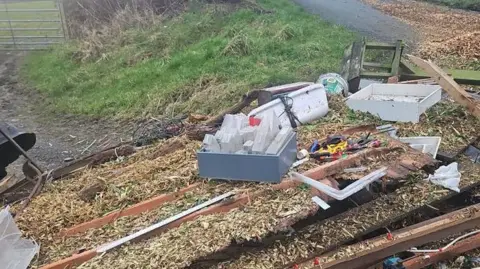 Westmorland and Furness Council Fly-tipped wasted which has been dumped at the side of a road. There a number of plastic cans, aerosol cans, snapped wooden planks, garden items and a large wooden sideboard. 