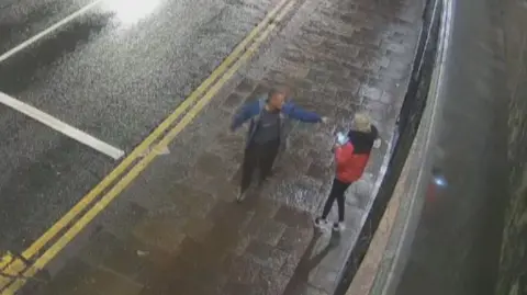 COPFS CCTV footage of Michael Harvey arguing with Carol O'Reilly on a street. He is pointing in her face, She is wearing a red and black jacket and he is wearing a blue jacket 