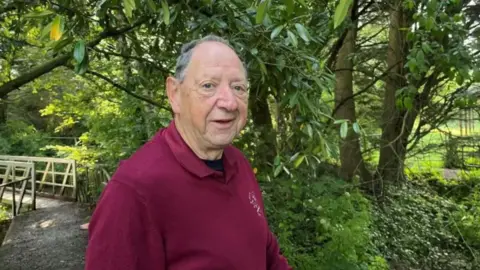 Jeremy Nettle looks into the camera wearing a dark red jumper. He is standing in the nature reserve with trees behind him.